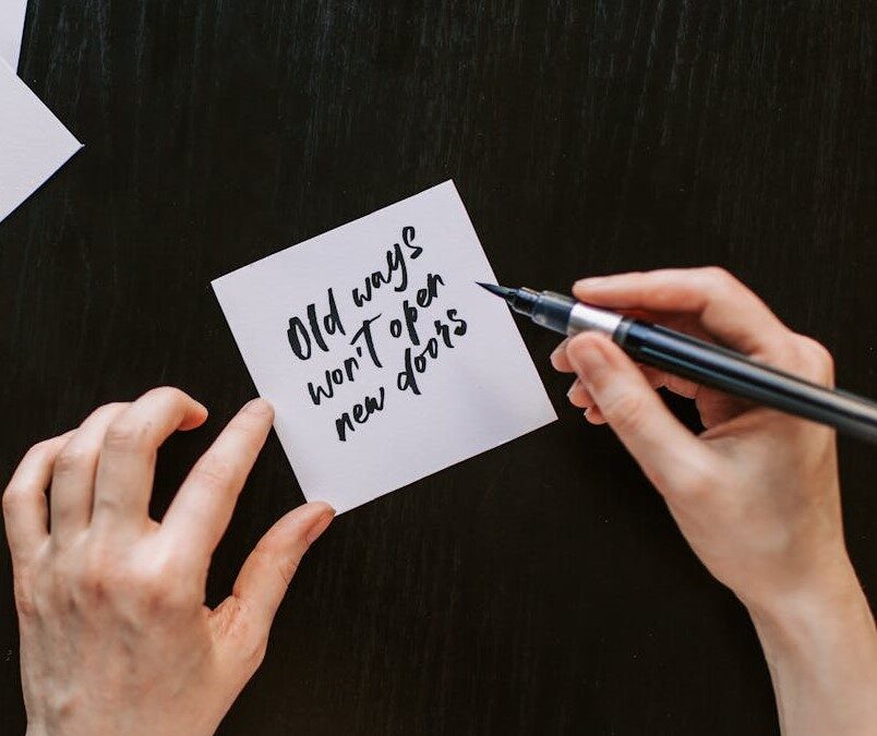 person writing on a note that says "Old ways won't open new doors." ; evolve as a leader